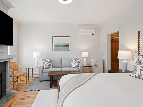 The image shows a cozy bedroom with a bed, sofa, TV, lamps, a fireplace, and a modern decor theme, featuring neutral tones and wooden accents.
