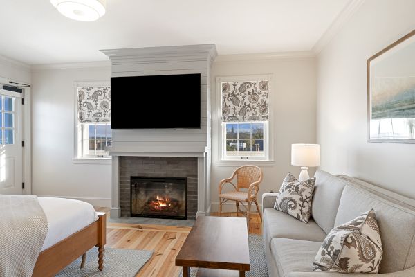 A cozy living room with a sofa, pillows, wooden table, fireplace, and TV. There’s a bed, wicker chair, lamp, and patterned curtains.