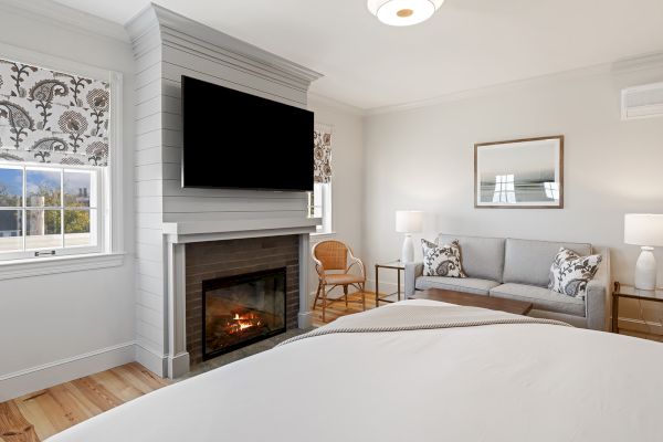 A cozy bedroom features a fireplace, TV, bed, sofa, and window with floral curtains, creating a warm and inviting atmosphere.