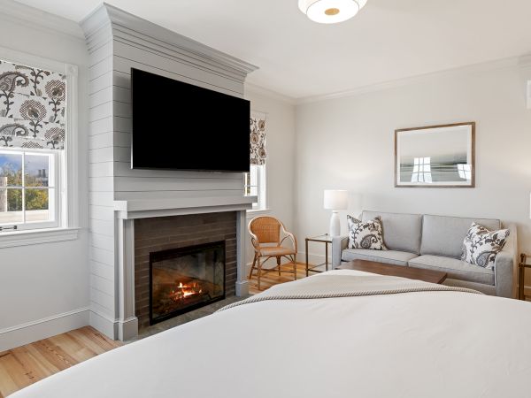 A cozy bedroom features a fireplace, TV, bed, sofa, and window with floral curtains, creating a warm and inviting atmosphere.