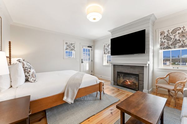 The image shows a cozy bedroom with a bed, fireplace, TV, and a chair, featuring neutral decor and wooden flooring.