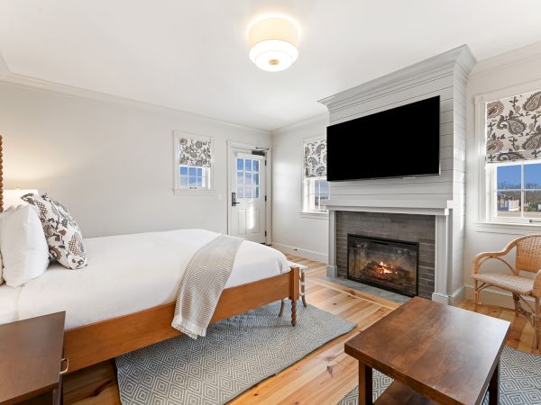 The image shows a cozy bedroom with a bed, fireplace, TV, and a chair, featuring neutral decor and wooden flooring.