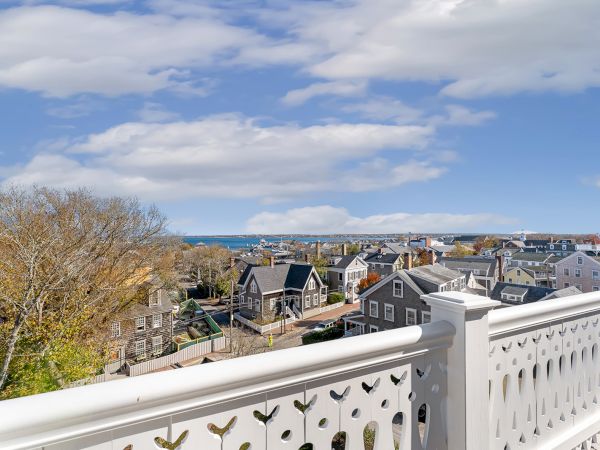 The image shows a scenic view of a coastal town with houses, trees, and distant ocean, seen from a balcony with a white railing.