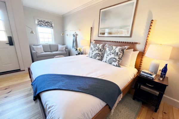 A cozy bedroom with a large bed, decorative pillows, and a blanket. There's a small couch, lamps, and a framed picture above the bed.