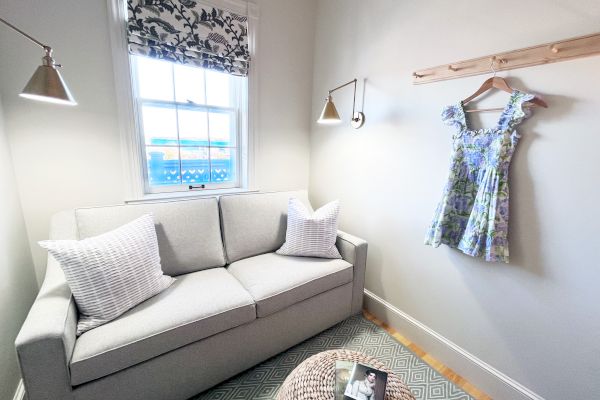 A cozy room with a gray sofa, a floral dress on the wall, two reading lamps, and a window with a view.