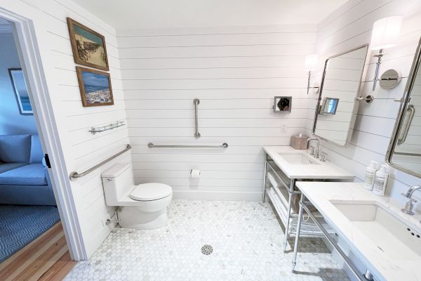 A white bathroom with a toilet, double sinks, and a mirror. The room has a shiplap wall and tiled floor, featuring grab bars for accessibility.