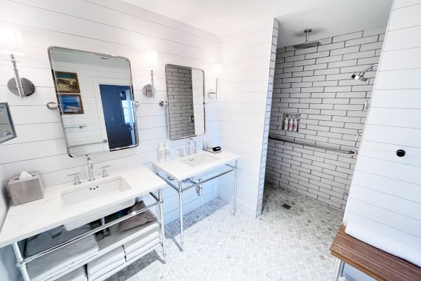 A modern bathroom with two sinks, mirrors, a walk-in shower with white subway tiles, and a wooden bench, all in a sleek design.