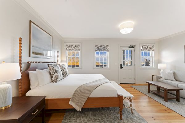 This image shows a well-lit bedroom with a large bed, side tables, and a sofa. The room has wooden floors and three windows with patterned shades.