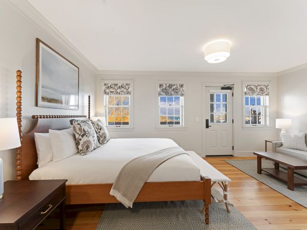 This image shows a well-lit bedroom with a large bed, side tables, and a sofa. The room has wooden floors and three windows with patterned shades.
