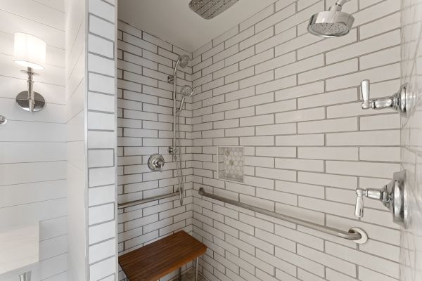 A tiled shower with two showerheads, a grab bar, and a wooden bench. There's a light fixture visible on the left.