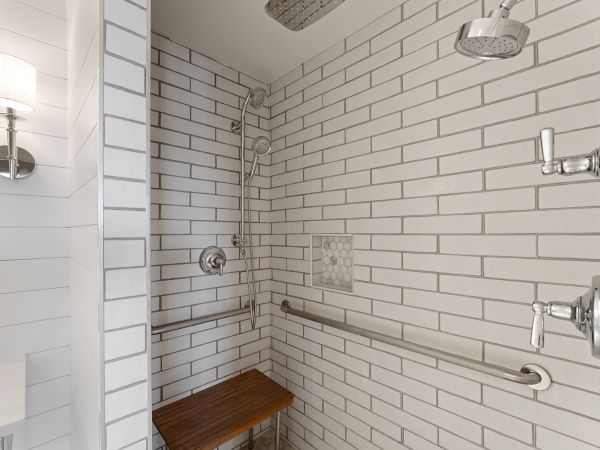 A tiled shower with two showerheads, a grab bar, and a wooden bench. There's a light fixture visible on the left.