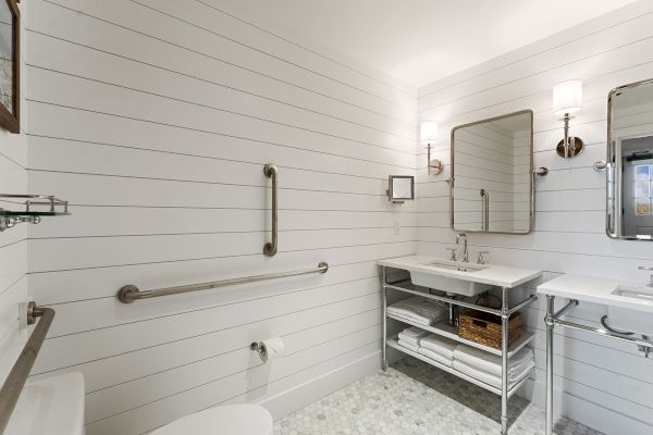 A modern bathroom with white shiplap walls, dual mirrors, a metal shelf, towels, two sinks, and a toilet. Wall-mounted lights add elegance.