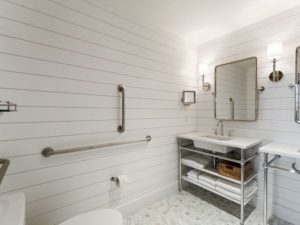 A modern bathroom with white shiplap walls, dual mirrors, a metal shelf, towels, two sinks, and a toilet. Wall-mounted lights add elegance.
