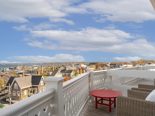 A rooftop deck with a white railing, red table, and wicker chairs overlooking a cityscape under a cloudy sky.