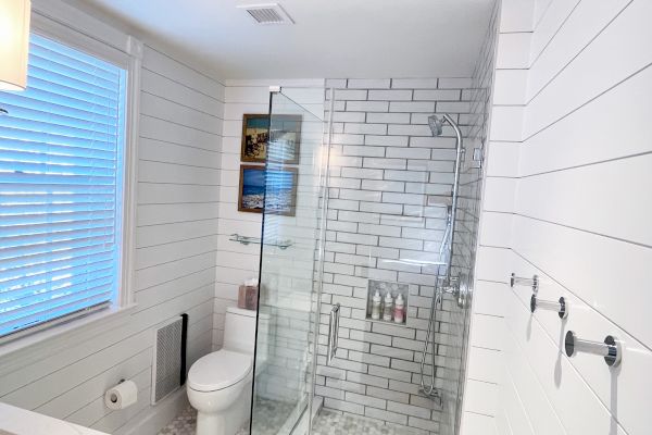 The image shows a modern bathroom with a glass shower, white subway tiles, a toilet, and wall art. It has a clean, minimalist design.