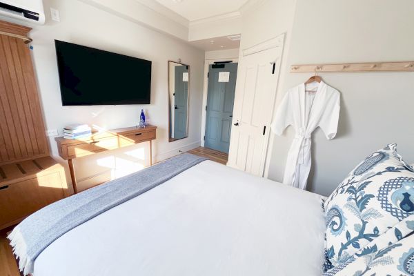 A bright bedroom with a bed, wall-mounted TV, desk, robe on a hook, and decorative pillows.