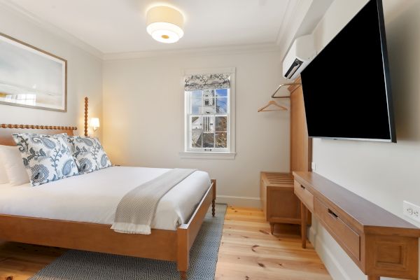 The image shows a cozy bedroom with a double bed, TV, window, and minimalist decor, featuring light walls and wooden furniture.