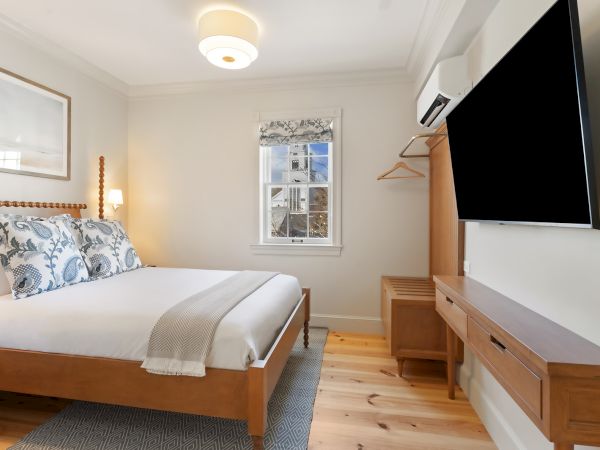 The image shows a cozy bedroom with a double bed, TV, window, and minimalist decor, featuring light walls and wooden furniture.