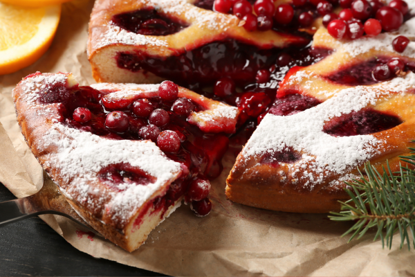 A lattice-topped pie with red berries is dusted with powdered sugar, accompanied by orange slices and evergreen sprigs.