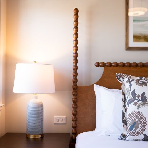 A cozy bedroom scene with a wooden bed, decorative pillows, a bedside lamp, and wall art. Natural light coming through a window enhances the space.