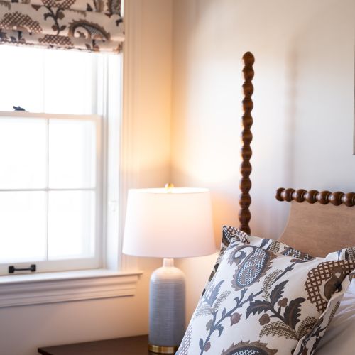 A cozy bedroom scene with a patterned pillow, elegant lamp, and decorated window shade beside a wood-framed bed against a light-colored wall.