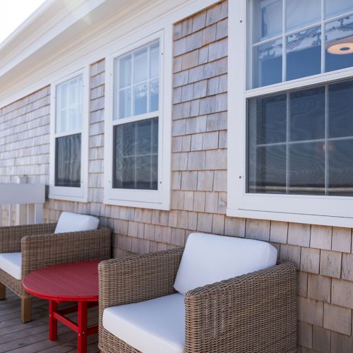 The image shows a patio with two wicker chairs with white cushions and a small red table, set against a house with wooden shingle siding.