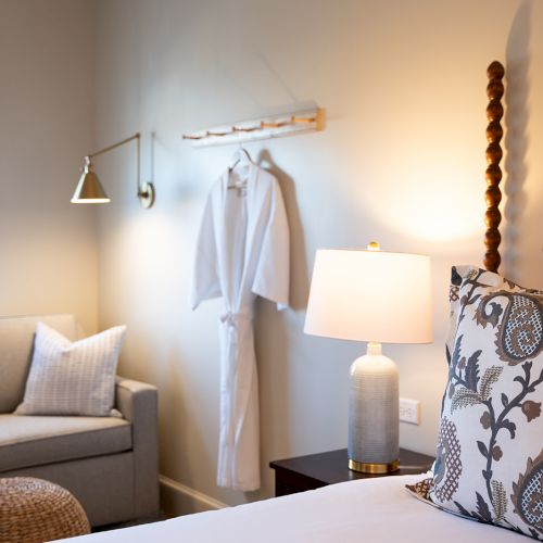 A cozy bedroom features a sofa, a lamp, a robe hanging on the wall, and decorative pillows with a floral pattern on the bed.