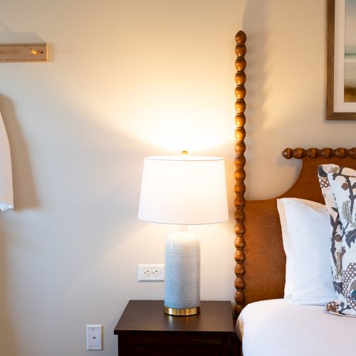 A cozy bedroom with a wooden bed, patterned pillow, white lamp on a nightstand, wall art, and a robe hanging on a hook.