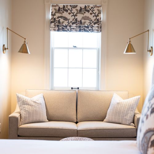 A cozy room features a gray sofa with pillows, wall-mounted lamps, and a window with floral curtains, viewed from a bed with patterned pillows.