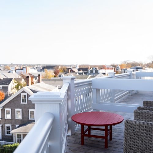 A rooftop deck with wicker chairs, a red table, white railings, overlooking a residential neighborhood and distant landscape.