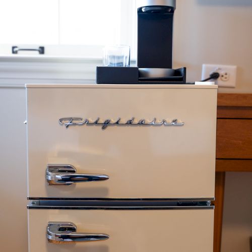 A vintage-style Frigidaire mini fridge with two doors is topped with a coffee maker beside a window and a wooden table.