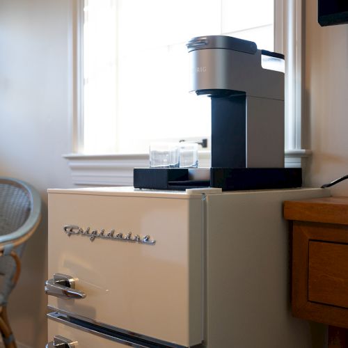 A retro-style Frigidaire mini fridge with a coffee maker on top, next to a wicker chair and wooden table by the window.