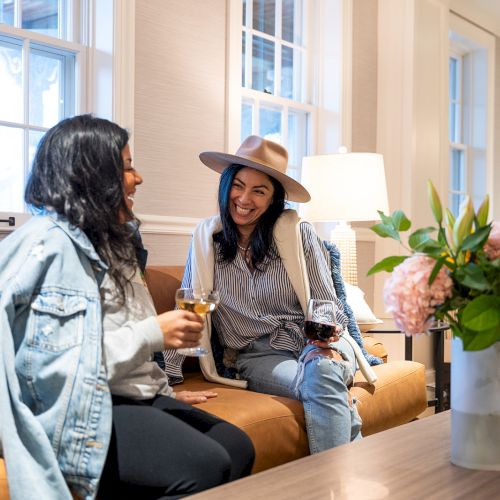 Two people are sitting on a couch, enjoying drinks and chatting. A vase with flowers is on the table in a cozy room.