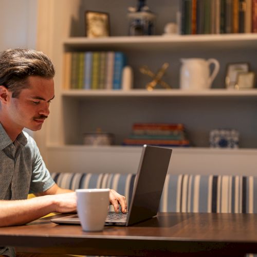 A man is working on a laptop at a table with a mug nearby, set in a cozy room with bookshelves in the background.