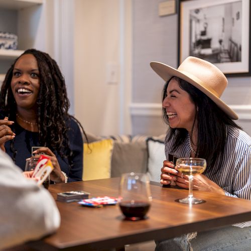 Three people are sitting at a table, enjoying drinks and playing cards, laughing and talking in a cozy room.