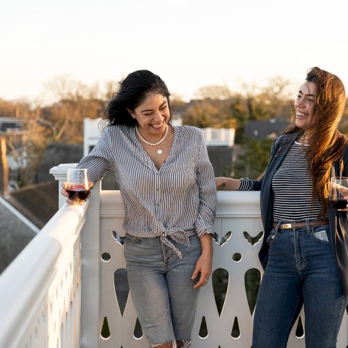 Two people are smiling on a balcony, holding drinks, with houses and trees in the background as the sun sets.