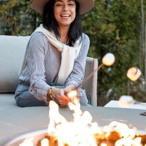 A person in a hat smiles while roasting marshmallows over a firepit outdoors, surrounded by greenery.