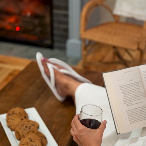 A person relaxes by a fireplace with a book, cookies on a plate, and a glass of wine, giving a cozy and comfortable atmosphere.