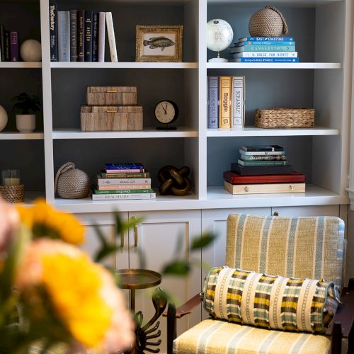 A cozy room with a bookshelf filled with books, decorative items, and plants. Two striped chairs and a small round table complete the scene.