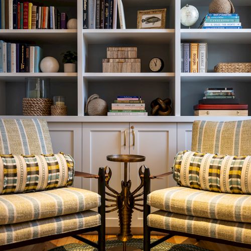 Two striped armchairs with patterned cushions face a small table in front of a bookshelf filled with books, decor, and a clock.