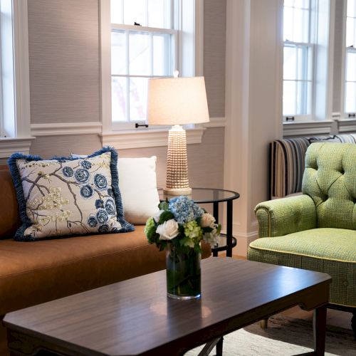 A cozy living room with a brown sofa, green armchair, floral pillows, table with a flower vase, and a lamp by the windows.