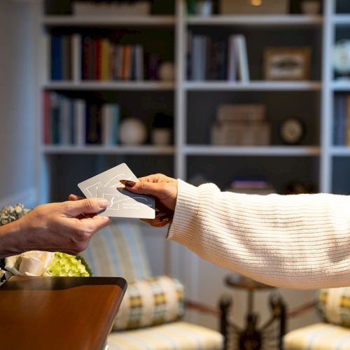 Two hands exchanging cards in front of a cozy bookshelf setting, suggesting a check-in or exchange scenario.