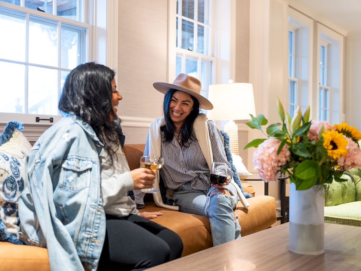 Two people are sitting on a couch, smiling and holding drinks, with a vase of flowers on the table nearby.