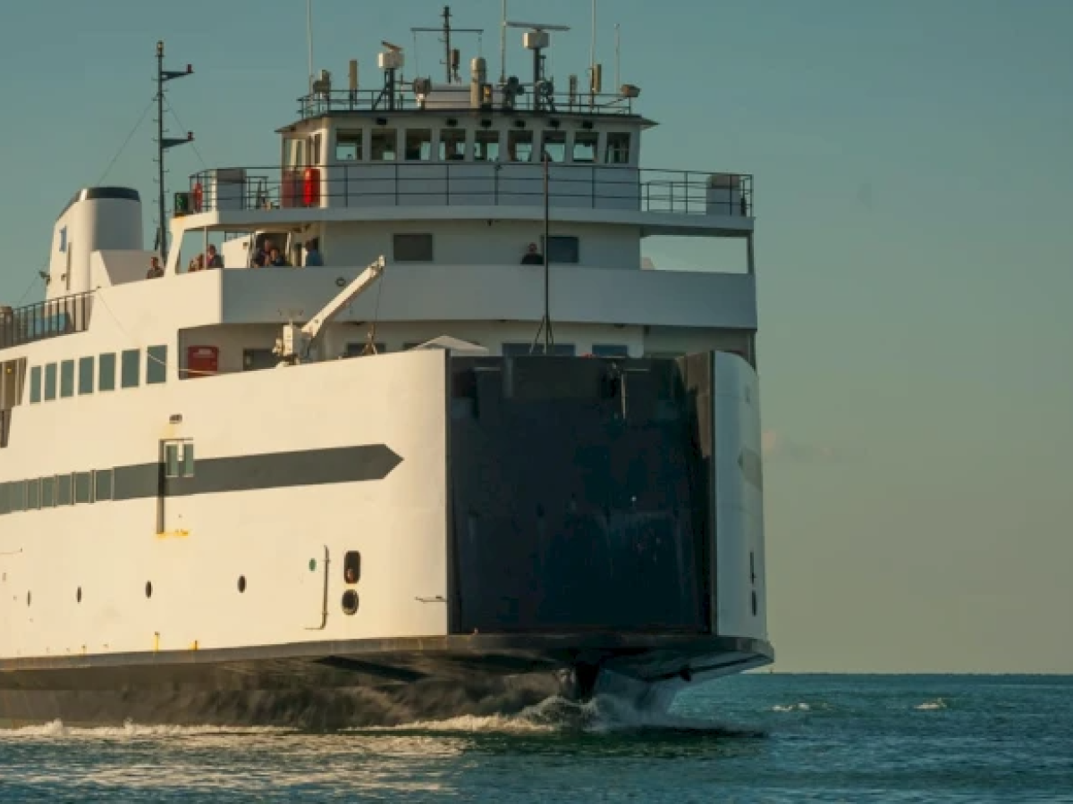 The image shows a large ferry moving through calm waters under a clear sky, with several people visible on the deck.