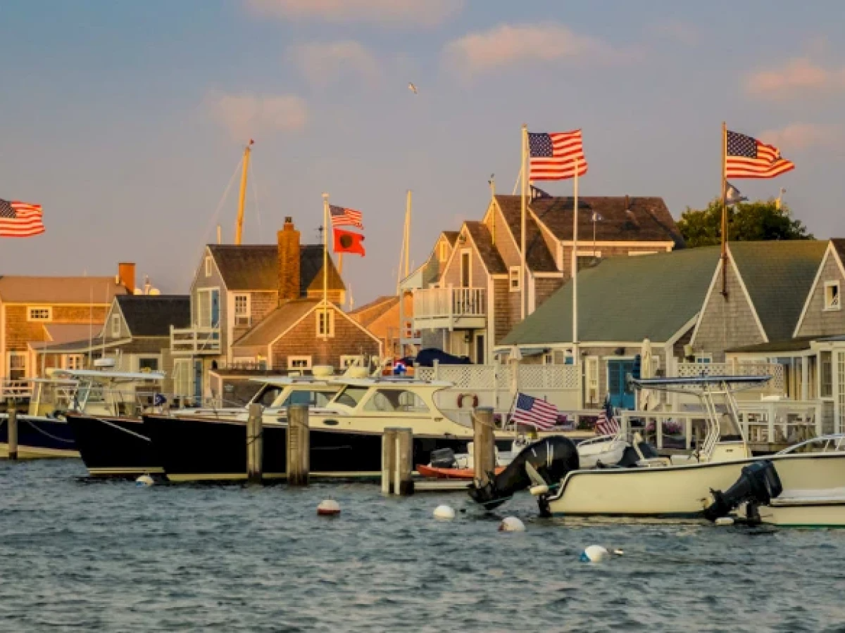 Coastal scene with boats docked by charming houses displaying American flags under a blue sky.
