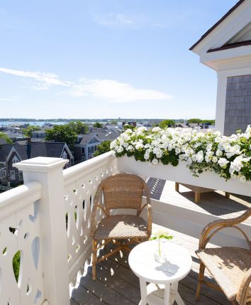 A sunny balcony with wicker chairs, a white railing, hanging white flowers, and a distant waterfront view beyond rooftops.