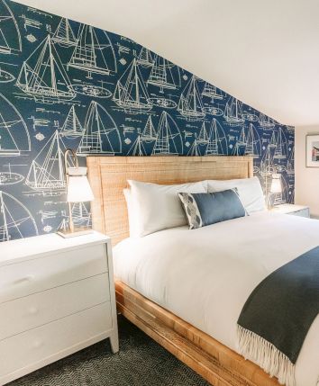 A bright bedroom with a wooden bed, nautical sailboat wallpaper, two windows with blinds, and blue accents by the chair and bedside lamp.