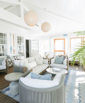 Sunlit modern patio with white sofas, wicker hanging lamps, nautical wheel wall decor, and lush palm leaves.