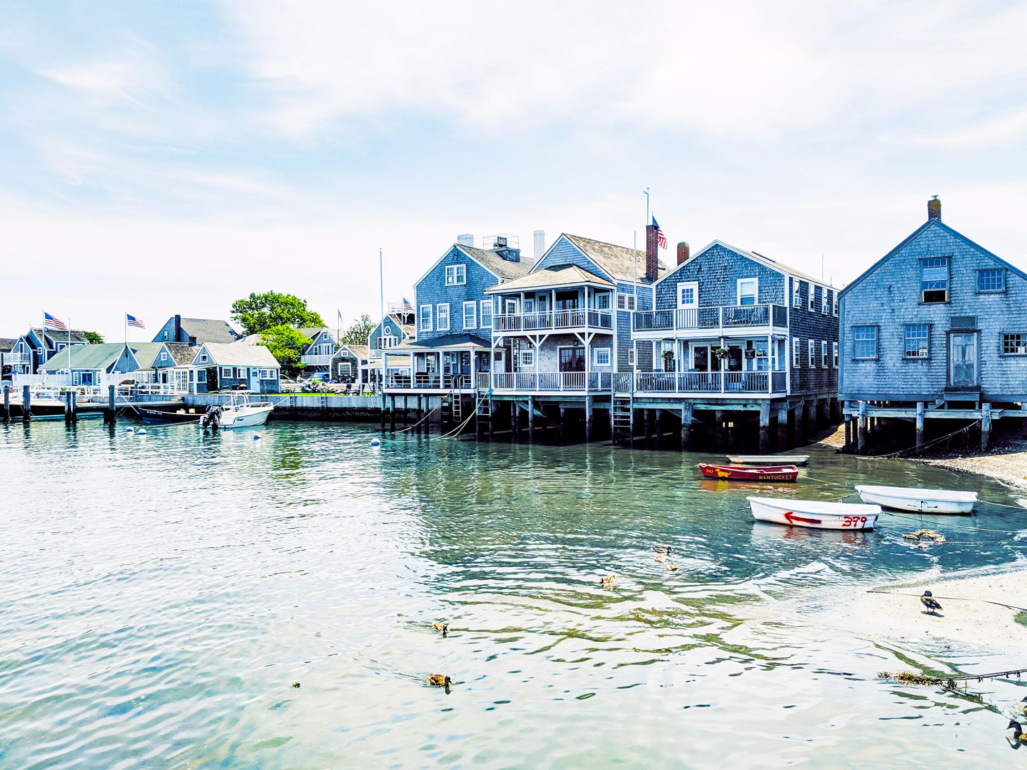 Coastal scene with wooden houses on stilts, small boats in the water, and ducks swimming nearby. Bright and tranquil atmosphere.