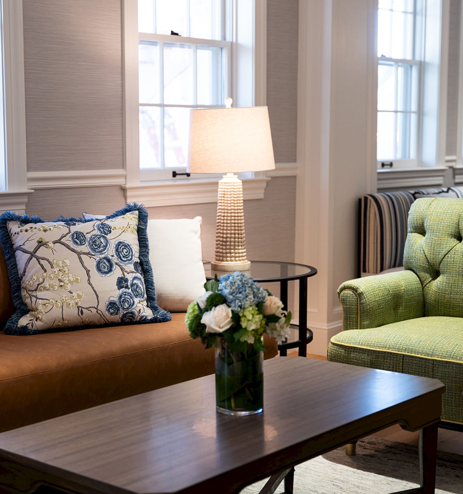 A cozy living room with a brown sofa, green armchair, table with flowers, a lamp, and decorative pillows by windows.